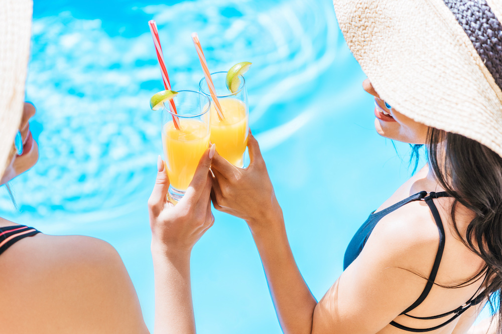 cropped shot of young women in swimwear and hats holding glasses with refreshing beverages at pool